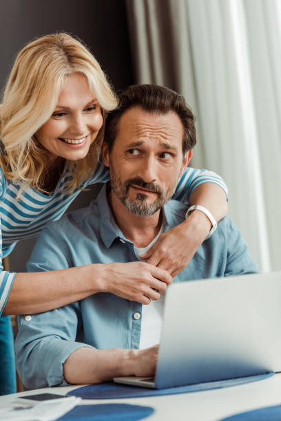 istock Selective focus of smiling woman embracing confused husband using laptop in kitchen 1250456727