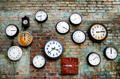 A lot of vintage clocks of different sizes hanging on a grunge brick wall. Round and square clocks show different time. Some of the clocks have no hands.