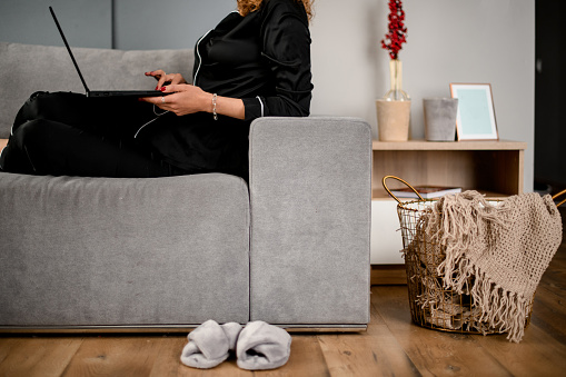 woman in black pajamas with laptop lies on the sofa. next to the floor are slippers and wicker basket with plaid inside