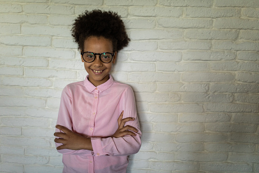 Portrait of cute mixed race girl with Afro hair, standing in front of white brick wall with arms crossed, looking at camera and smiling