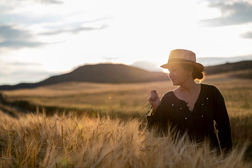 Young woman farmer with straw hat is walking in wheat field at sunset