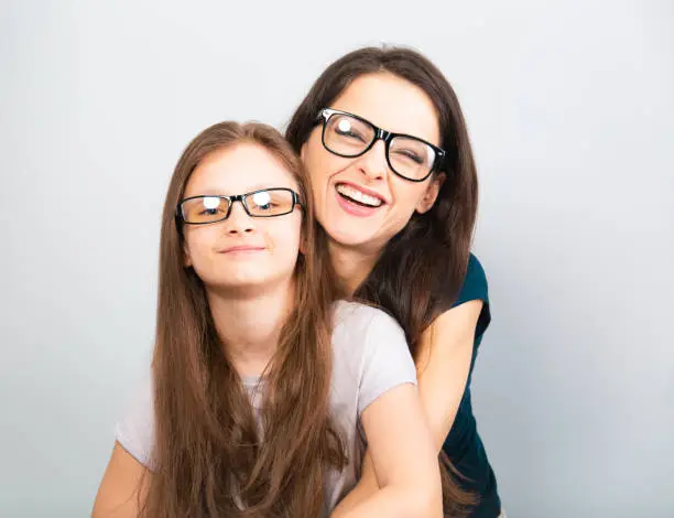 Happy young casual mother and smiling kid in fashion glasses hugging on light blue background with empty copy space. Closeup studio portrait
