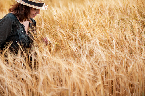 Young woman farmer with straw hat is walking in wheat field at sunset