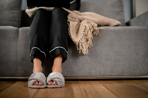 front view on legs of woman which sitting on sofa with knitted blanket in black silk pajama and grey slippers