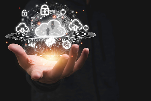 A close-up of a human hand extending outward, holding a glowing 3D representation of cloud technology symbols, including a cloud icon, lock symbols, and graphs, set against a dark background.
