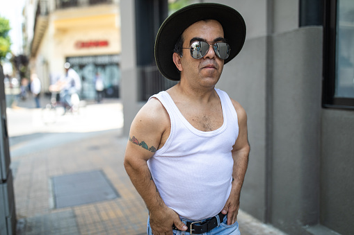 Cool attitude man with dwarfism, with tattoo on arm, wearing jeans and undershirt, with hat and sunglasses on walking down city streets in Buenos Aires