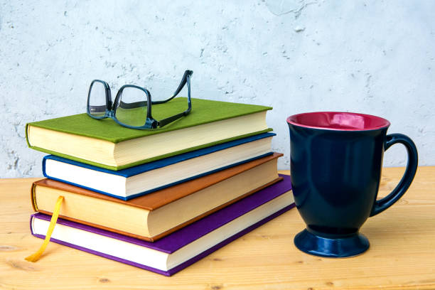 stack of books on wooden table with eyeglasses and cup on grey background
