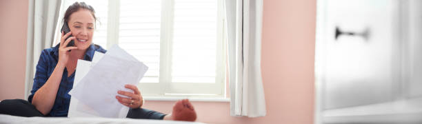 Young woman sat on her bed talking on the phone happily sorting her financial matters