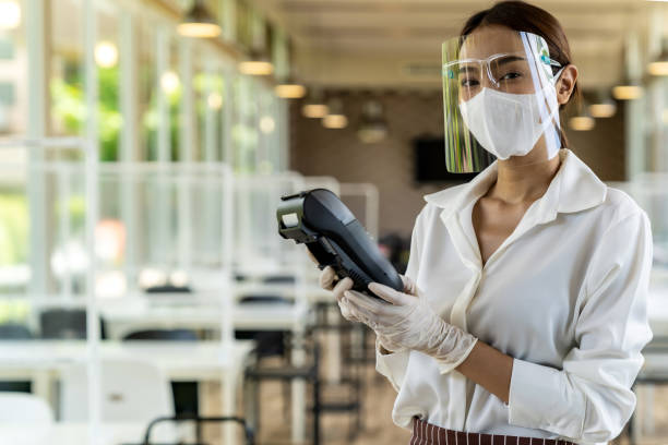 Waitress with face mask hold credit card reader. Portrait attractive asian waitress wear face mask and face shield hold credit card reader for contactless payment with indoor restaurant background. New normal restaurant contactless payment concept. asian cashier stock pictures, royalty-free photos & images