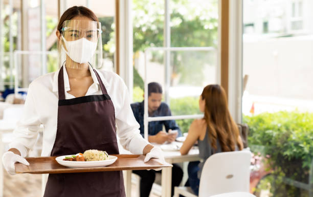 Asian waitress serving food new normal. Portrait attractive asian waitress wear face mask and face shield holding food tray to serving meal to customer with custome in background. New normal restaurant concept. reopening photos stock pictures, royalty-free photos & images