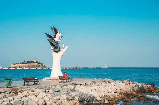 Kusadasi, Aydin Province, Turkey - September 9, 2019: Man Sitting on bench near Hand Of Peace Monument On Waterfront In Sunny Summer Day. View Of Hand Sculpture At Aegean Coast, Turkey.