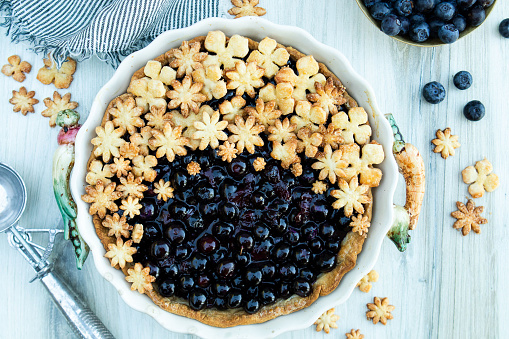 This flaylay photo of a fresh blueberry pie shows glistening blueberries in a flaky pie crust.  The flower cutouts on the top and surrounding the pie give a lovely playfulness to this pie.  The pie sits in a white scalloped pie dish, on a pale wooden surface.  There are additional blueberries and flowers surrounding the pie, along with an ice cream scoop and striped napkin.