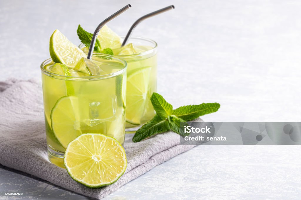 Two glasses with iced green matcha tea with lime, ice, fresh mint and metal drinking straws on light grey backdrop. Two glasses with iced green matcha tea with lime, ice, fresh mint and metal drinking straws on grey napkin on light grey backdrop. Healthy food, No Plastic, Zero Waste concepts. Copy space for text. Drink Stock Photo