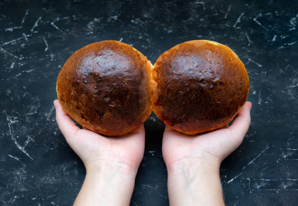 Female hands holding two freshly baked golden brown buns against a textured black background. Top view, selective focus, studio shot.