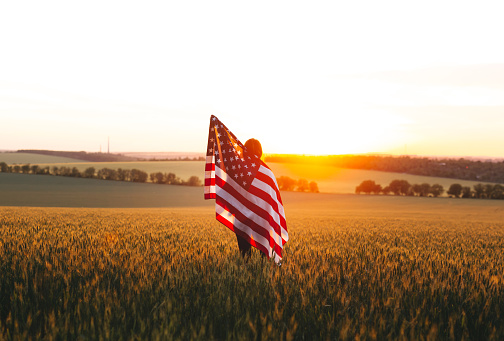 Woman with the American flag  in a wheat field at sunset. Adventure lifestyle. Independence Day, 4th of July.