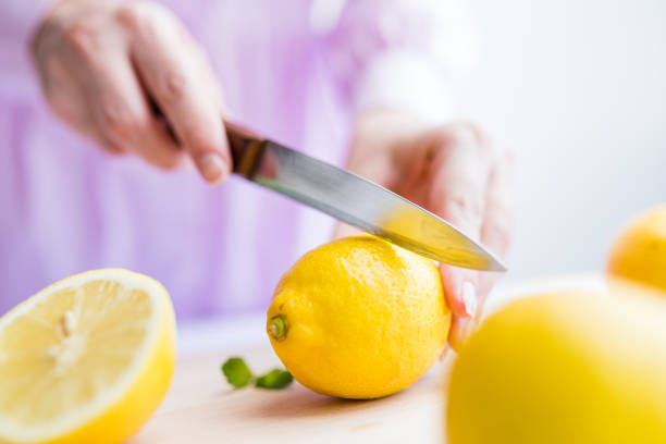 Female hands slicing lemon on half with knife on cutting board.