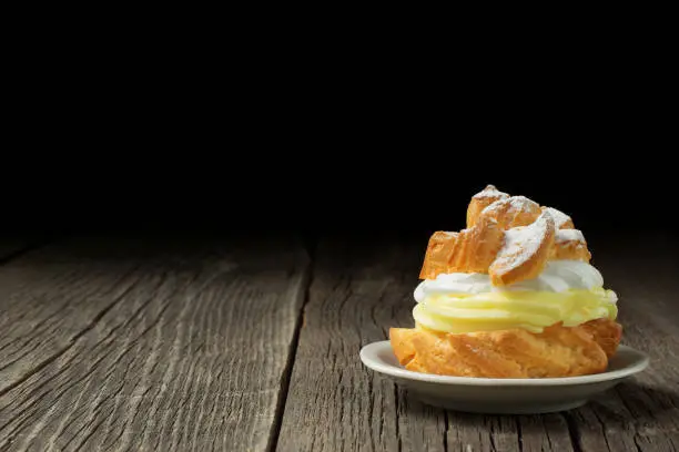 A profiterole, decorated with a dusting of powdered sugar and filled with an abundant amount of custard, is lying on a small ceramic saucer, on rustic weathered wood planks. A lot of copy space.