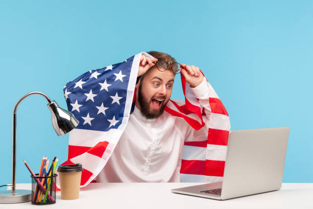 Amazed, extremely happy businessman sitting wrapped in American flag, watching live patriotic program on laptop Amazed, extremely happy businessman sitting wrapped in American flag, watching live patriotic program on laptop at workplace and shouting for joy with surprised shocked expression. indoor studio shot patriotic man stock pictures, royalty-free photos & images