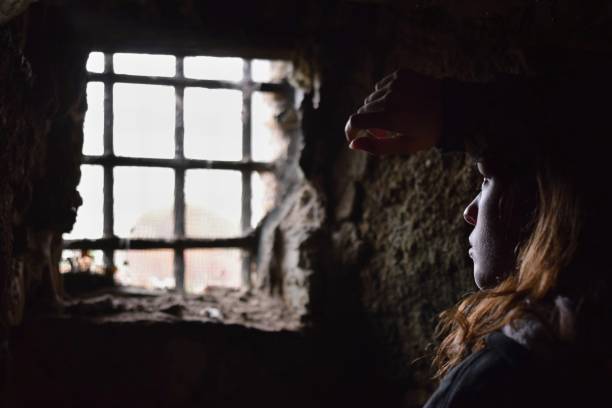 Young woman captive or prisoner watching desperately out from the window of her underground cell Young woman captive or prisoner watching desperately out from the window of her underground cell dungeon medieval prison prison cell stock pictures, royalty-free photos & images
