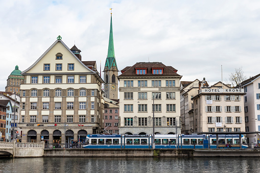 Zurich, Switzerland - November 2019: Predigerkirche Church Clock Tower seen between buildings across the Linmat River, featuring public transport tram