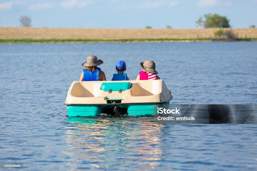 Rear View of Three Children In Paddle Boat on Small Lake Rear view of three children in a paddle boat on a small lake. Two girls are on either side of their little brother, who is sitting in the middle. Pedal Boat Stock Photo