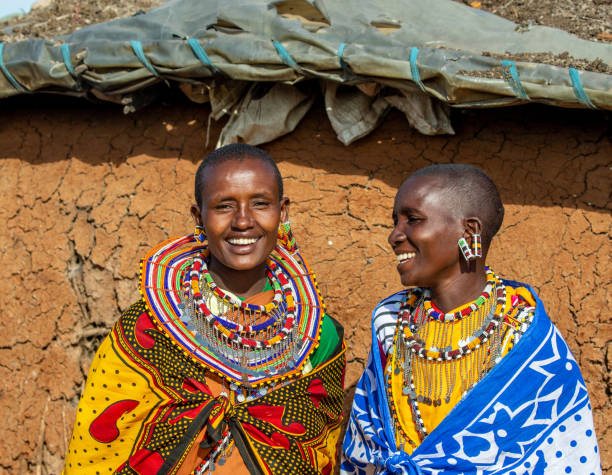 Two Masai women in traditional dress are talking to each other against the wall of a traditional house. stock photo