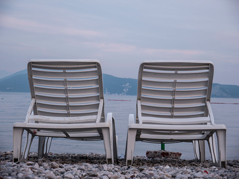 Two open empty white plastic chaise lounge laid out on sea side pebbles and mountains on background at the end of summer season side view