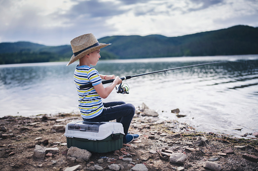 Side view of little boy with straw hat fishing in beautiful mountain lake.