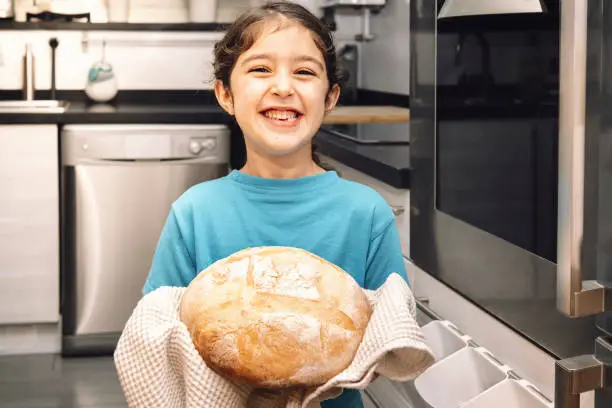 smiling little girl holding a loaf of freshly baked rustic bread in the kitchen, concept of healthy eating at home