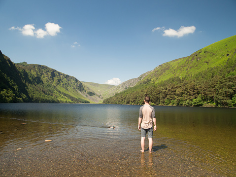 A young man in shorts and a t-shirt stands in the shallow water of the Glendalough Upper Lake. He looks out at the breathtakingly green Irish Hills. The sky is clear blue with only a few white clouds.