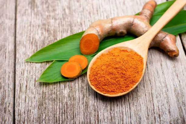 Turmeric powder in wooden spoon and tumeric root with slice and green leaf isolated on rustic wood table background. Top view.