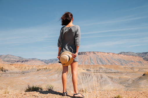 Fashionable girl with hat on a desert mountain top wearing grey dress. Woman back standing on the top of mountain and beautiful sky.