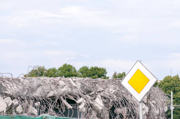 A heap of bricks, wood, and shattered windows marks the remains of a demolished building, with debris spread across bare earth as nearby homes and trees stand untouched in the distance