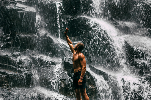 A man of athletic build at the waterfall. A man travels the world. Man at the waterfall, view from the back. Travel to Bali Indonesia. Back view of a man standing by waterfalls with arms outstretched