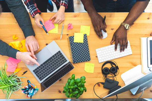 Multiracial coworkers in small home office top view and close up on hands and computer - Startup business or friends sharing a project - Cluttered office desk with people at work
