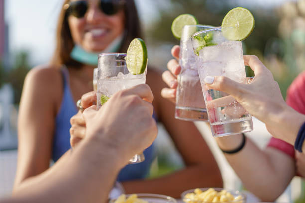 Friends drinking cocktails in an outdoors snack bar restaurant in the summer wearing face mask on to be protected from coronavirus - Happy people cheering with mojito and having fun stock photo