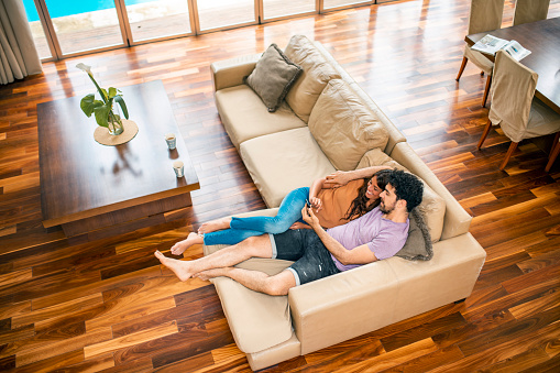 High angle view of mixed race and Hispanic couple in 20s and 30s reclining on sofa and checking smart phone together in modern home with hardwood floor.