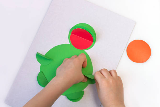 istock Concept of DIY and children's creativity. Step by step instruction: how to make applique frog from paper. Step 4 child fingers gluing paper paws. Creative activities with child during quarantine 1250622859