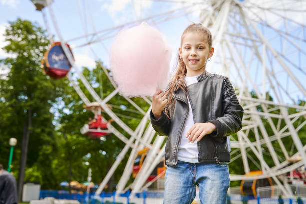 Portrait of a child with sweet cotton candy. A little girl on the background of ferris wheel is eating candy-floss summer sunny day Portrait of a child with sweet cotton candy. A little girl on the background of ferris wheel is eating candy-floss. child cotton candy stock pictures, royalty-free photos & images