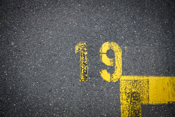 Number 19, yellow color thermoplastic road marking paint on the asphalt close up. Marked parking space ground background, delineated by road surface markings. No person space for text