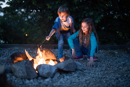 Happy little girl and her small brother enjoying by the campfire in the evening at the backyard. Copy space.