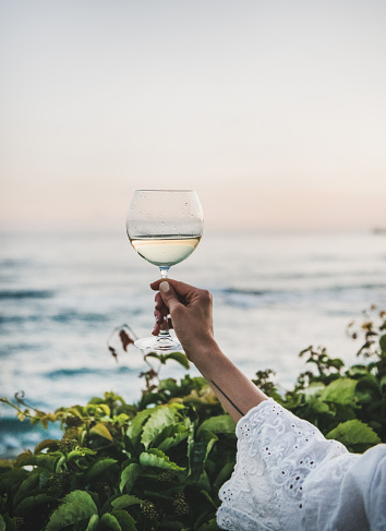 Womans hand in white lace dress sleeve holding glass of white wine with beautiful sunset colors and sea horizon at background. Summer picnic and refreshment drink concept