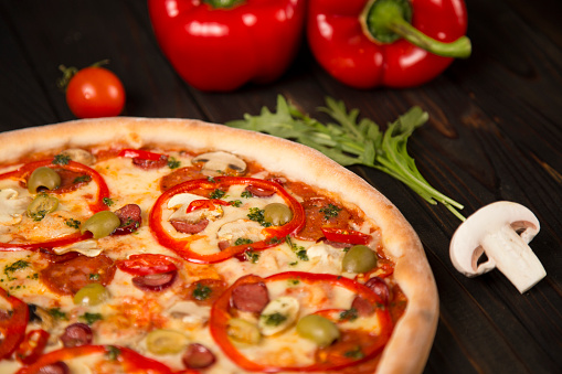 Side view of pizza with bell pepper, green olives, smoked sausages, mozzarella cheese, pepperoni, salami, chili pepper, champignons on wooden board with two bell peppers and herbs on background