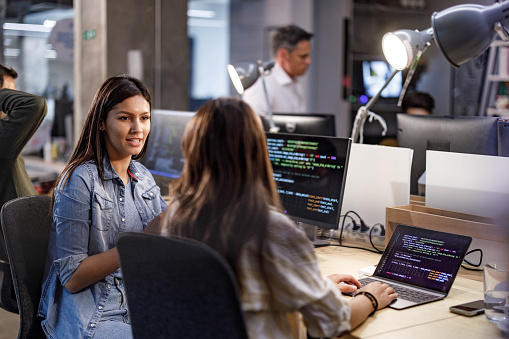 Smiling female programmer communicating with her colleague while working on computer codes in the office.