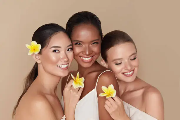 Photo of Diversity. Natural Beauty Portrait. Multi-Ethnic Women With Tropical Flowers Standing Together. Asian, Mixed Race And Caucasian Models With Nude Makeup And Healthy Skin Holding Plumeria.