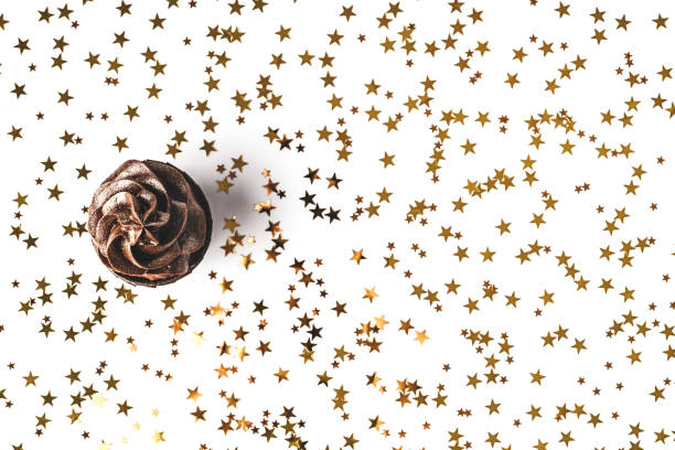 istock Chocolate cupcake with chocolate icing and sprinkled gold sparkles on white background with golden star shaped confetti. Holiday concept. Flat lay style. Copy space. 1250650815