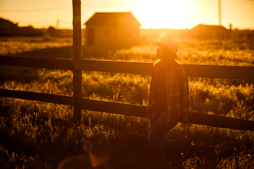 woman in hat and plaid shirt standing near a fence at sunset in the village. walks in the countryside. farming.