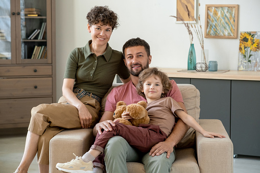 Happy young man with cute little son and his pretty wife sitting in large soft comfortable armchair in the center of living-room at home