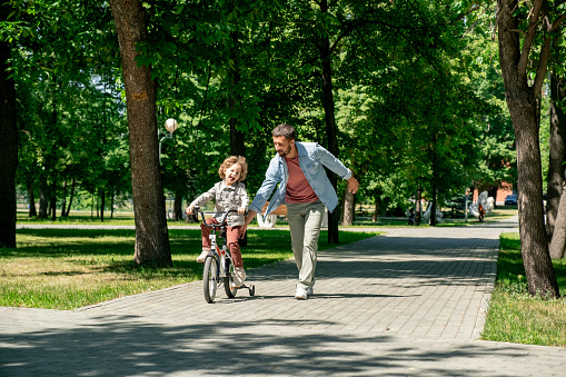 Joyful little boy riding bicycle with his father running near by along road surrounded by green lawns and trees in public park in summer