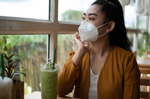 Joven mujer de Asia sentada y poniéndose una máscara médica para protegerse de las enfermedades respiratorias transmitidas por el virus que miró el té verde en un vaso en el café photo